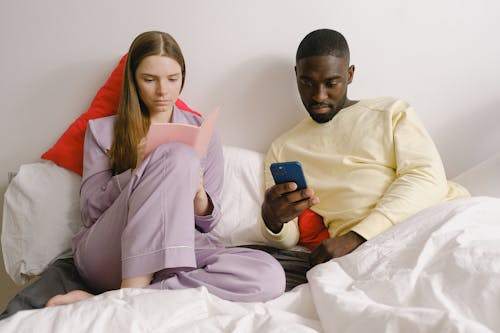 Free Serious woman taking notes in notebook near focused African American man while resting on comfortable bed in light bedroom at home Stock Photo