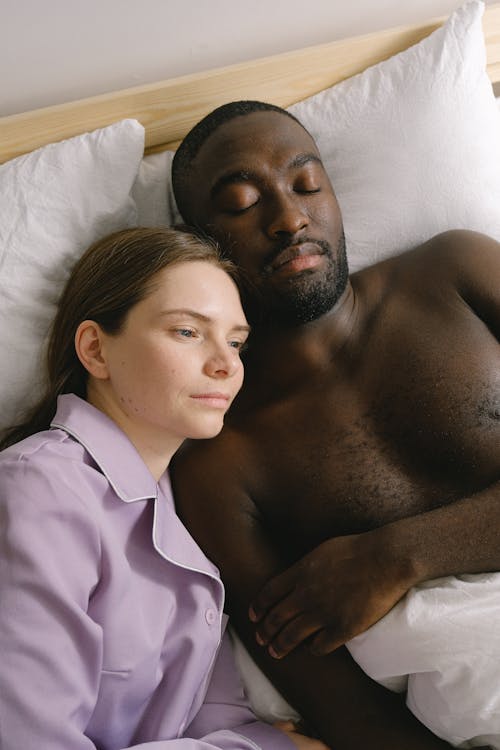 Free From above of calm woman in sleepwear near sleeping African American man lying in bed in light bedroom at home Stock Photo