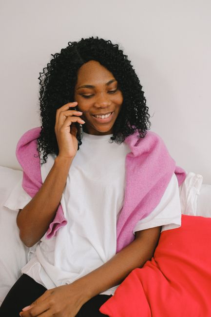 Content African American female having conversation on cellphone while sitting on comfortable bed with pillows in light bedroom at home