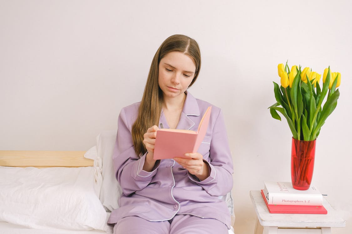 Focused female in nightwear writing in notepad while sitting between bed and chair with tulips and books in bedroom