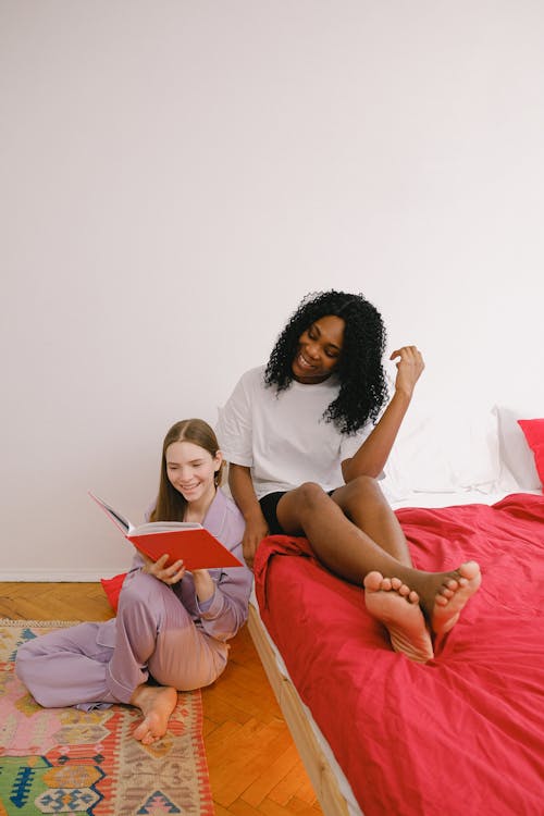 Free Full body of cheerful barefoot diverse female friends reading interesting book together in light bedroom with red blanket on bed at home Stock Photo