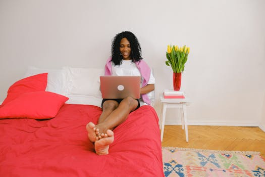 Smiling woman with curly hair using a laptop on a red bed in a bright bedroom with tulips.