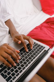 Crop unrecognizable African American person browsing internet on netbook while sitting on comfortable bed with colorful bedclothes in light bedroom at home