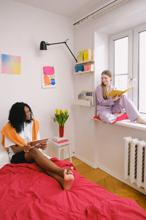 Free Full body of positive woman on windowsill with book in hands and African American female browsing tablet on bed while looking at each other in bedroom Stock Photo