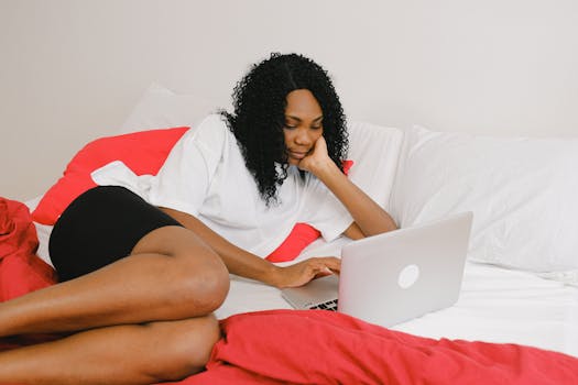 African American woman working on a laptop while relaxing in bed at home.