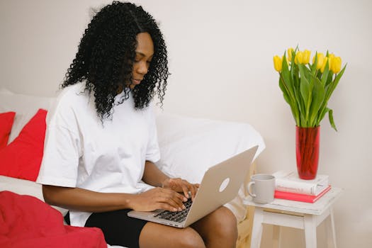 A woman types on her laptop in a comfortable bedroom with tulips and a coffee mug nearby.