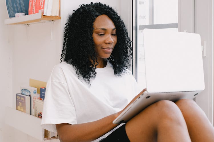 Happy Black Woman Working With Laptop Near Shelves With Books