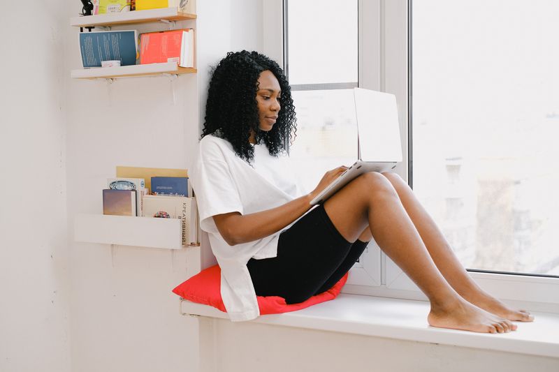 Black woman resting by a sunny window while using a laptop, representing accessible virtual wellness support