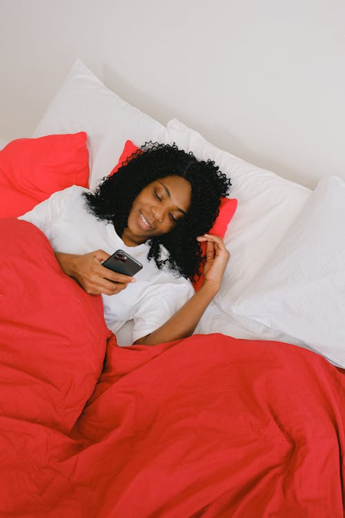 Free From above of positive young African American female checking mobile phone on soft white pillows in morning Stock Photo