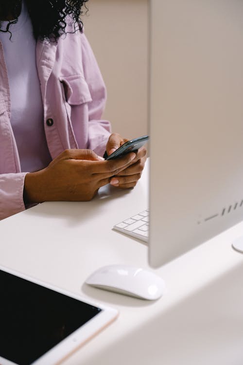 Crop unrecognizable African American female using mobile phone at white table with convenient computer in office