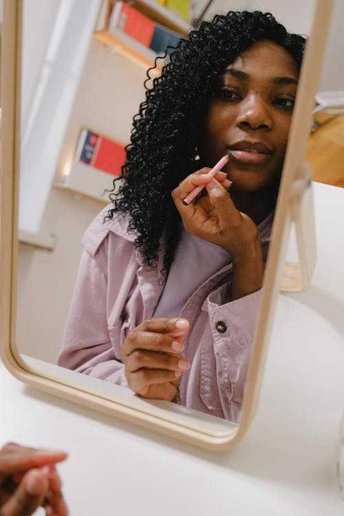 Pensive African American female with curly dark hair looking at mirror and applying lipstick while making makeup in light room