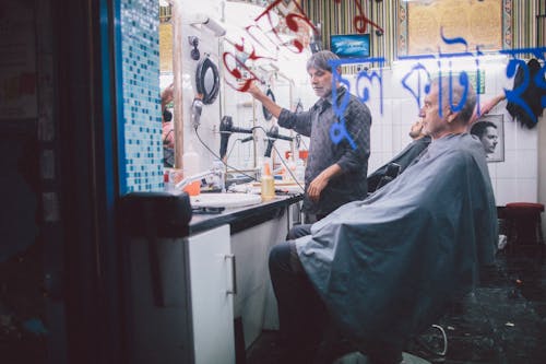 Elderly Man Sitting in the Chair with Blue Apron in Barbershop