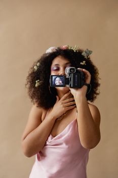 A woman uses a vintage camcorder, capturing her reflection, against a brown background.