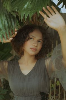 Dreamy young lady in trendy dress with raised hands looking away near plants with green leaves near fence in daytime
