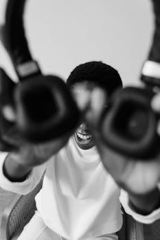 Black and white image of a person playfully holding headphones towards the camera with a joyful expression.