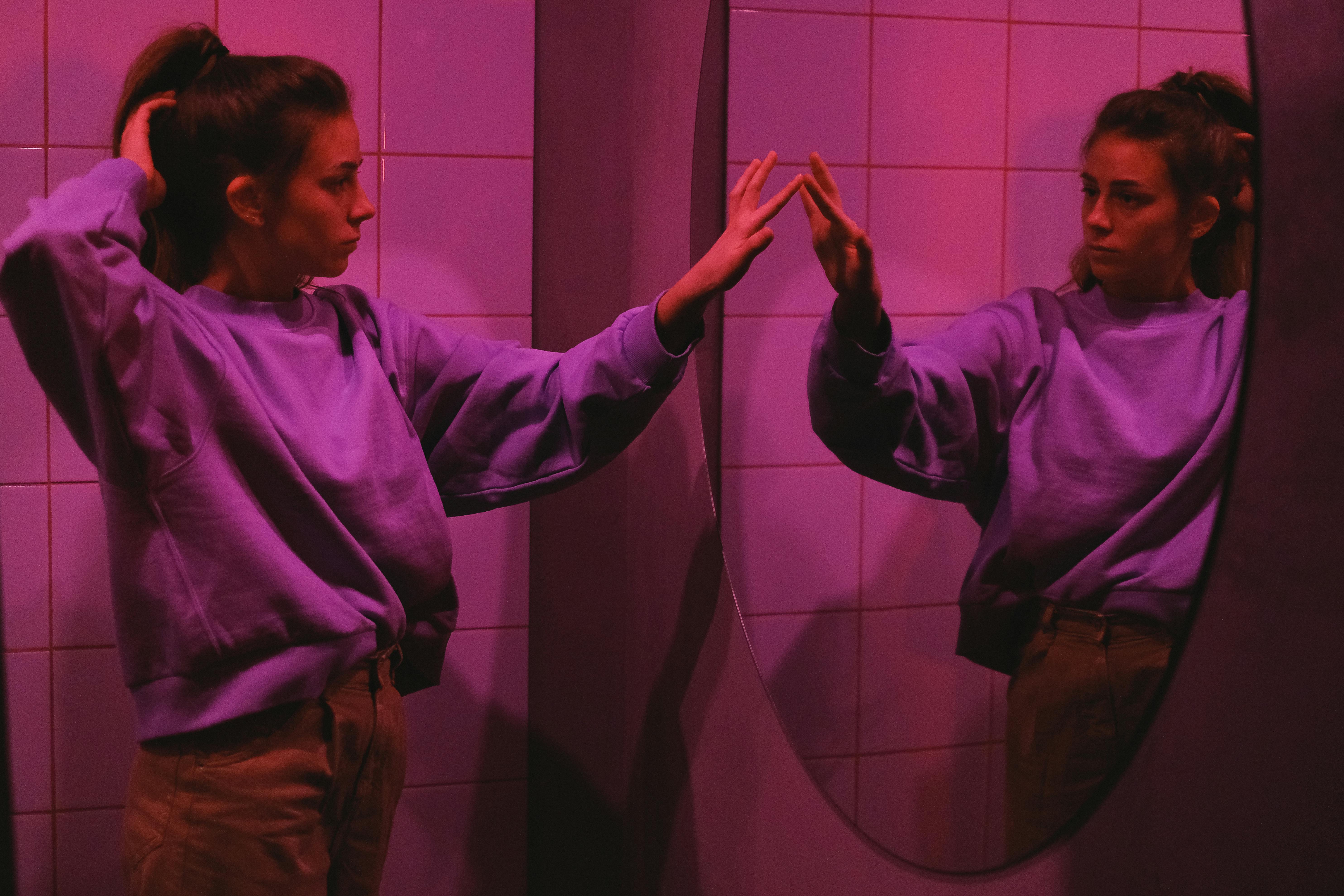 A young woman in a pink-lit room gazes at her reflection in the mirror, creating a moody and introspective atmosphere.