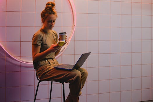 Young woman multitasks with phone and laptop in a trendy cafe with neon lighting.