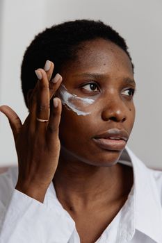 Black woman applying face cream indoors for skincare routine. Natural beauty and self-care focus.