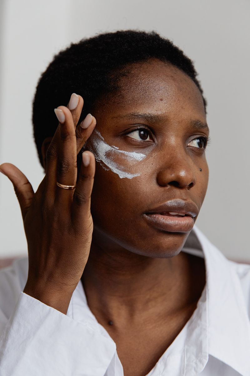 A Black woman applies skincare cream below her eye, focused on the cheek area where facial rosacea commonly appears.
