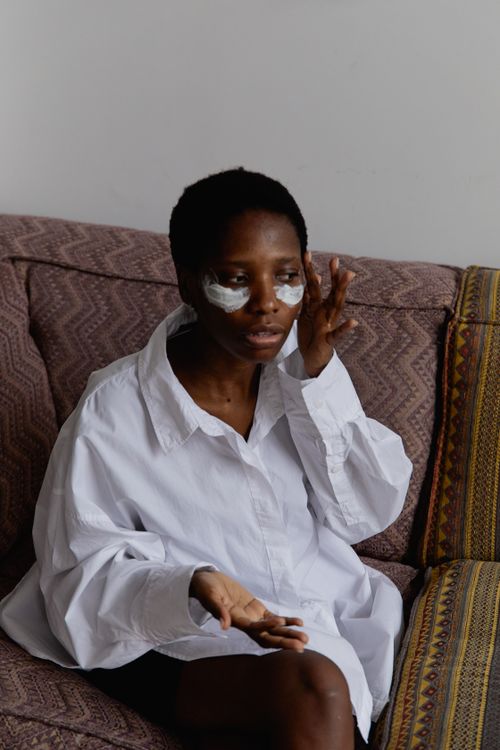 Free Black woman in a white shirt applying face cream while sitting on a sofa indoors. Stock Photo