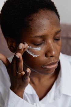 A close-up portrait of a woman with eyes closed, gently applying face cream indoors.
