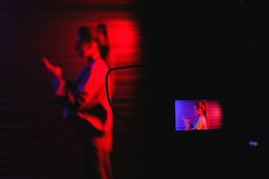 Side view of anonymous female standing and using smartphone while recording video on professional camera in dark room with neon lights