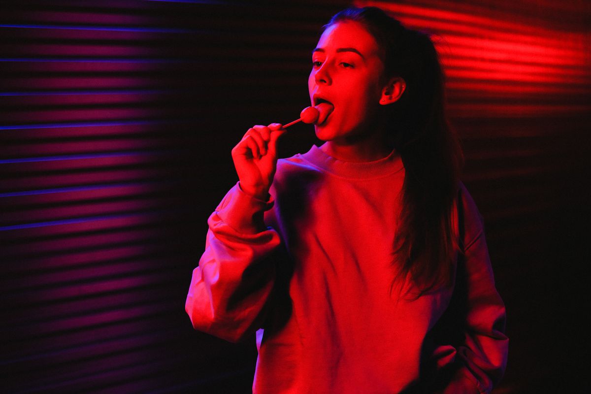 Portrait of a young woman enjoying a lollipop in a room with red lighting and shadow contrast.
