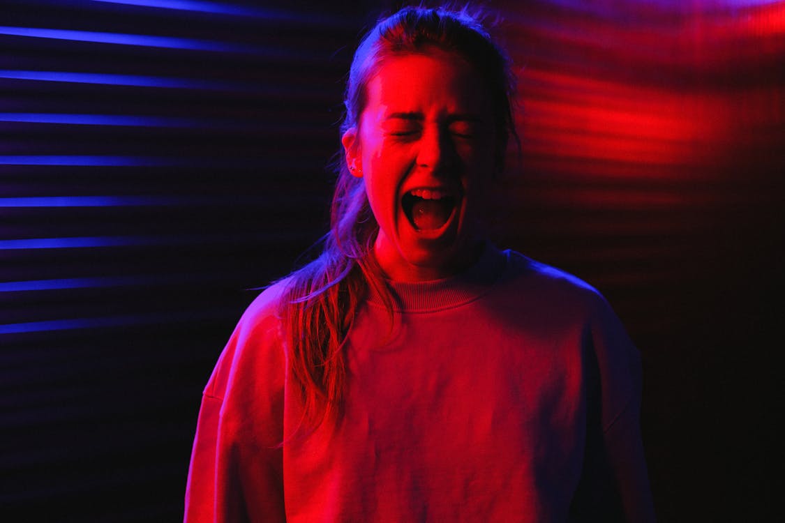 Frightened young woman standing with closed eyes and screaming in dark room with colorful neon lights
