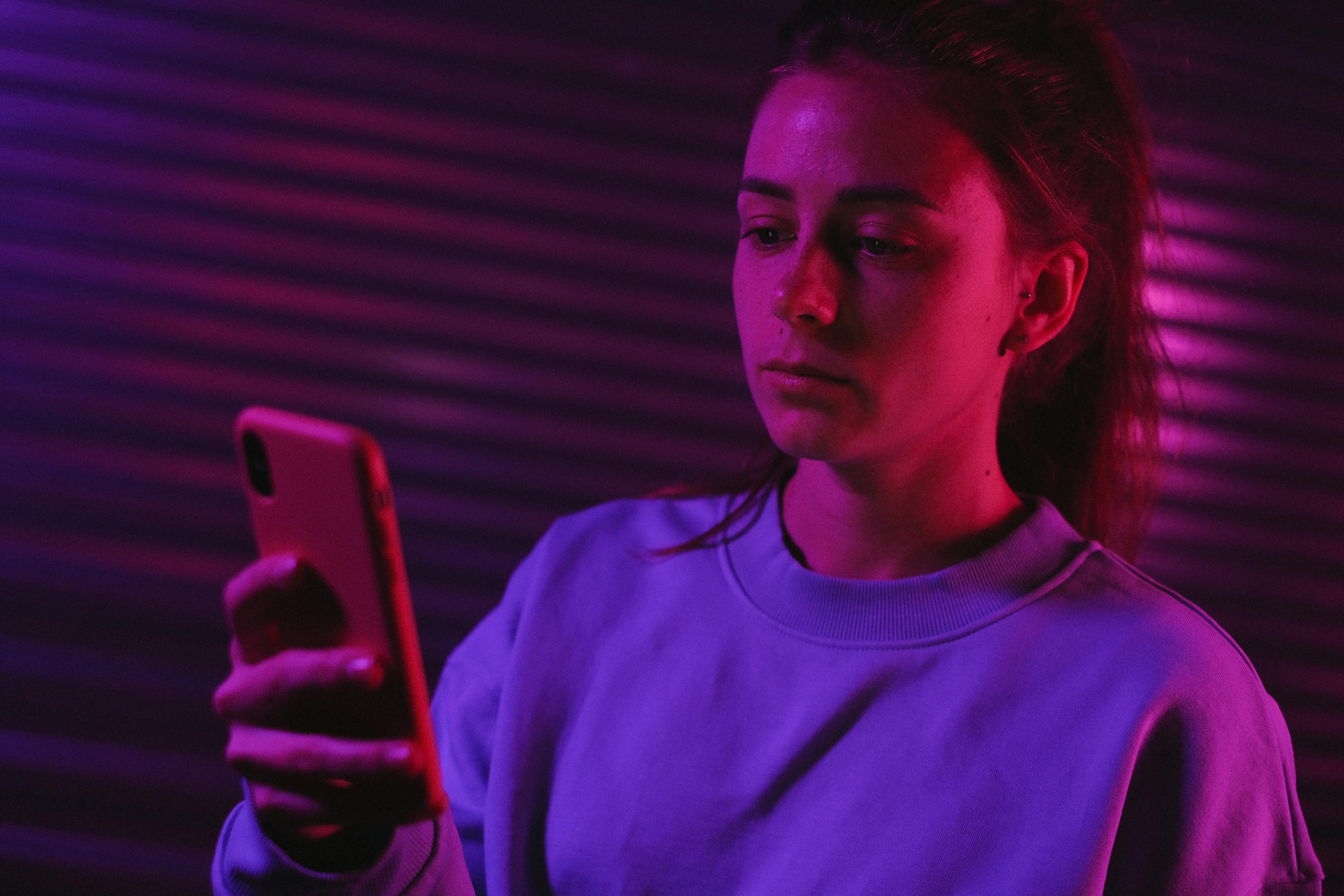 pensive female browsing smartphone in dark room with colorful lights