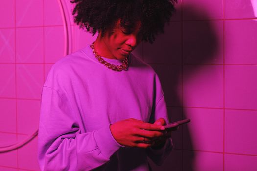 Stylish young woman with afro hair texting on phone in a modern pink-lit setting.