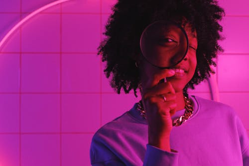 Woman in Purple Sweater Looking Through a Magnifying Glass Illuminated in Pink Light
