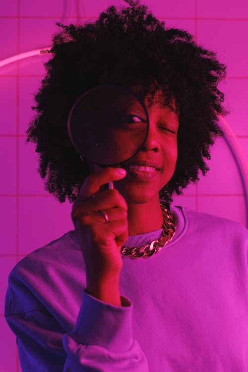 Positive African American woman with dark curly hair in casual clothes looking at magnifier in room with neon lights