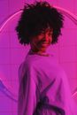 Side view of cheerful African American female teenager with Afro hairstyle looking at camera in pink light against ring lamp