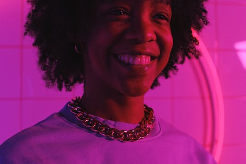 Crop smiling ethnic female teen in necklace with Afro hairstyle looking away in neon light against tiled wall