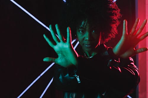 Attractive young African American female wearing trendy black leather jacket crossing arms and looking at camera while standing in illuminating neon lights in dark studio