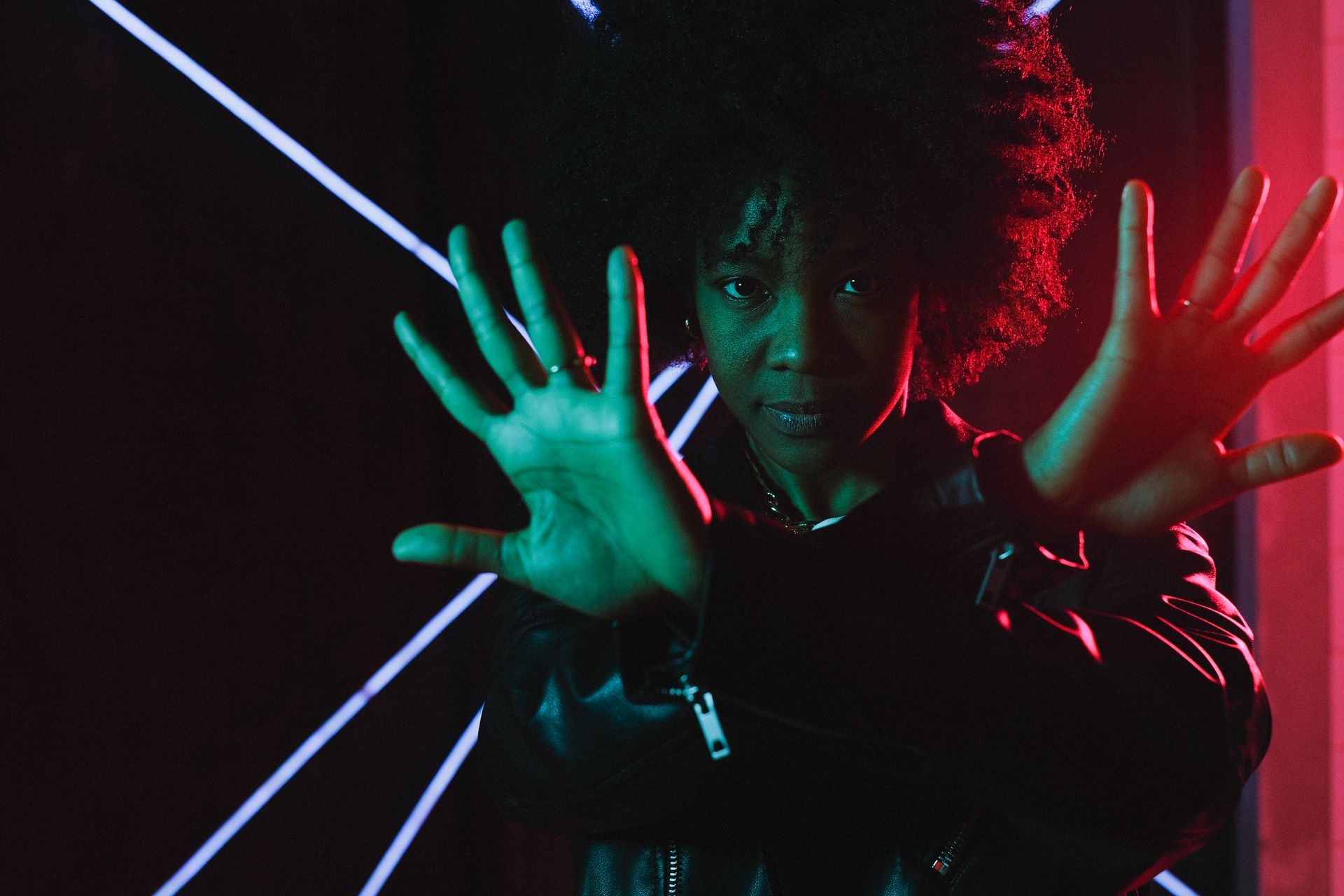Attractive young African American female wearing trendy black leather jacket crossing arms and looking at camera while standing in illuminating neon lights in dark studio