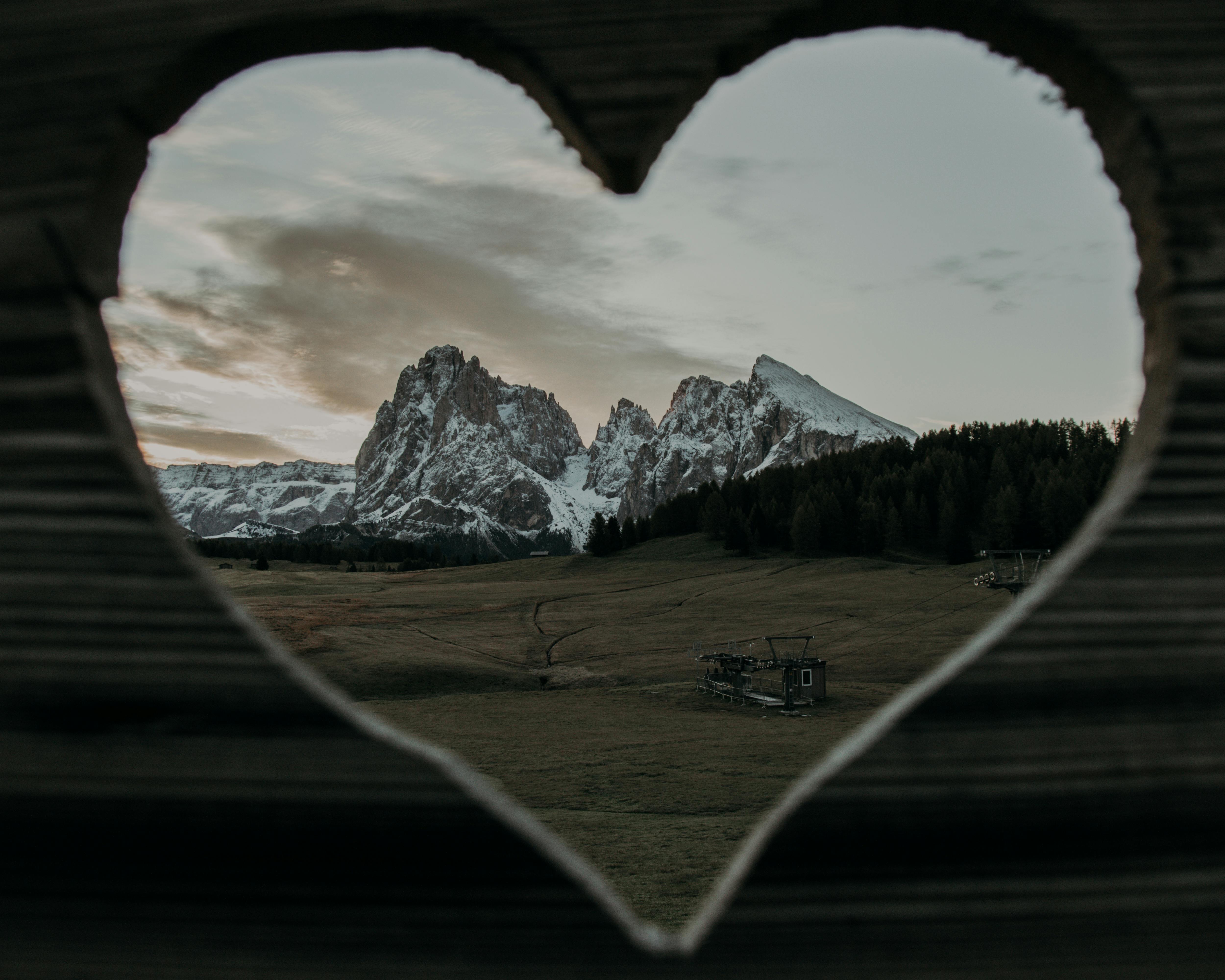 Through heart shaped hole of wooden fence on rocky cliffs covered with snow located on grassy field in rural area