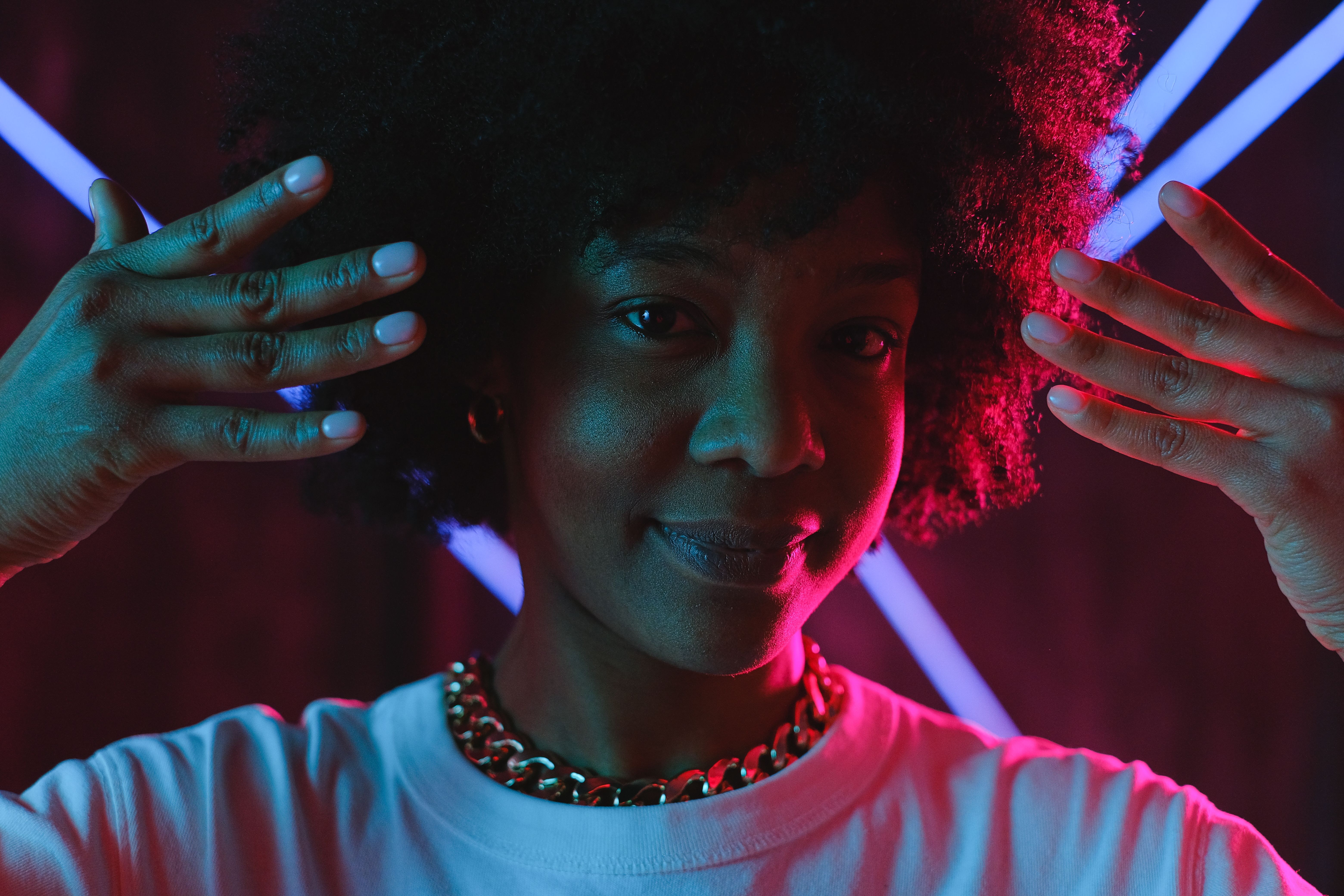 Smiling African American female in white shirt raising hands near pretty face and looking at camera while standing in neon lights in dark studio