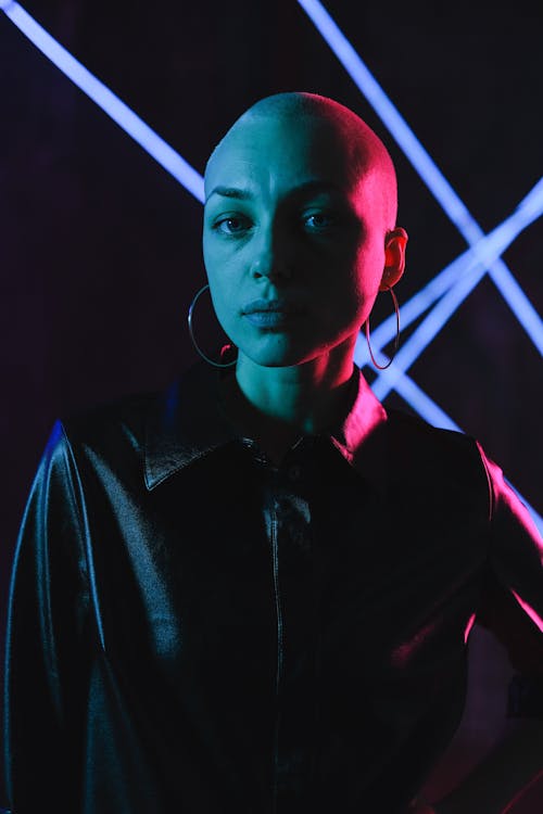 Emotionless young bald female in black leather shirt standing in dark studio in pink neon lights and looking at camera
