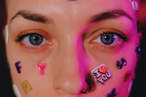 Crop calm female with nose piercing and letter stickers on face standing on dark background and looking at camera