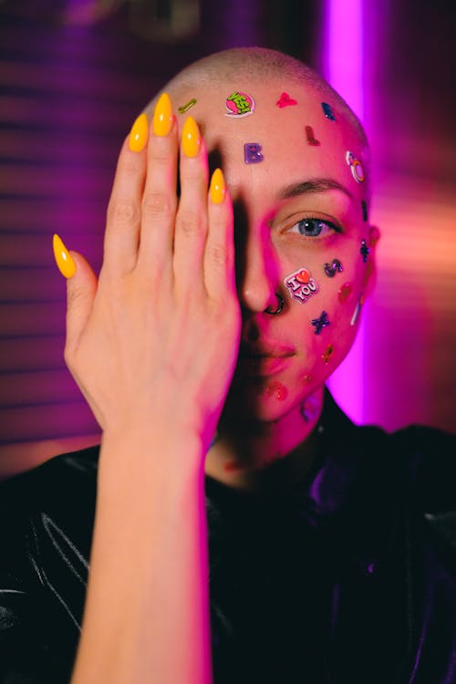 Emotionless bald female with small multicolored stickers on face covering half face with hand and looking at camera in dark studio