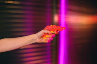 Crop anonymous female with manicured hand pointing toy gun against neon lights in dark studio