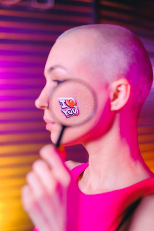 Young bald female with magnifying glass demonstrating sticker I Love You on face skin while standing in studio