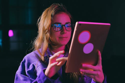 Concentrated young female in eyeglasses and casual outfit surfing modern tablet while standing in dark room with illumination during online work