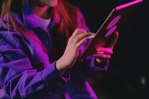 Crop unrecognizable female in casual clothes surfing internet on tablet while standing in dark room with illumination during online work