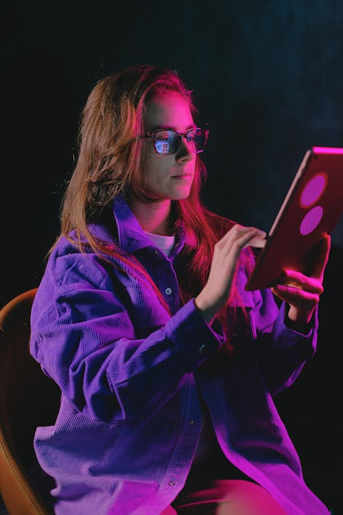Pensive female in eyeglasses surfing internet on modern tablet while sitting on black background in dark room with glowing illumination