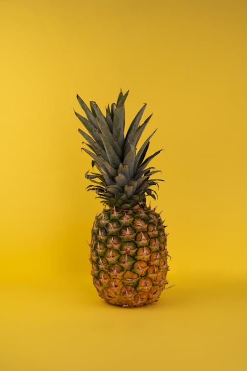 Whole fresh edible ripe pineapple fruit with green crown and brown skin placed on yellow background in modern light studio