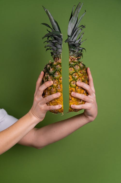 Crop anonymous person with halves of fresh pineapple with green leaves in hands standing on green background in light studio