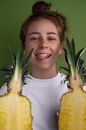 Positive female with halved ripe pineapple with yellow flesh looking away while standing with tongue out on green background in light studio