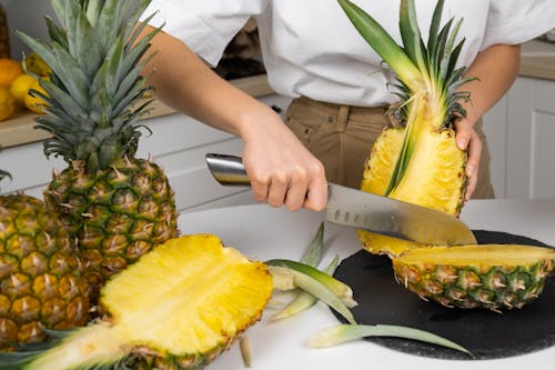 Crop anonymous cook cutting ripe pineapples with green leaves on table using big knife
